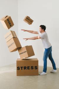 Image of a person wearing a white t-shirt and jeans pushing over cardboard boxes labeled as "stress,"problems," "anxiety," "deadline," "work, "breakup" to symbolize overcoming negative thoughts.